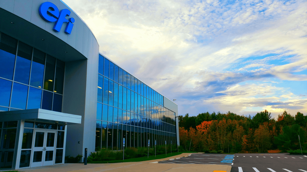 Modern building with large glass windows reflecting a vibrant sky. The "efi" logo is visible. Trees with autumn foliage line the background.